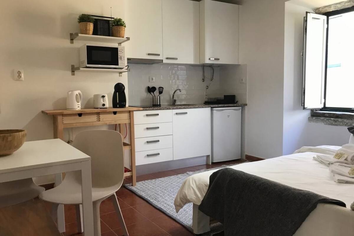 A compact kitchen with white cabinets, a microwave, and a coffee maker. There is a dining table with a chair and a bed with a blanket.
