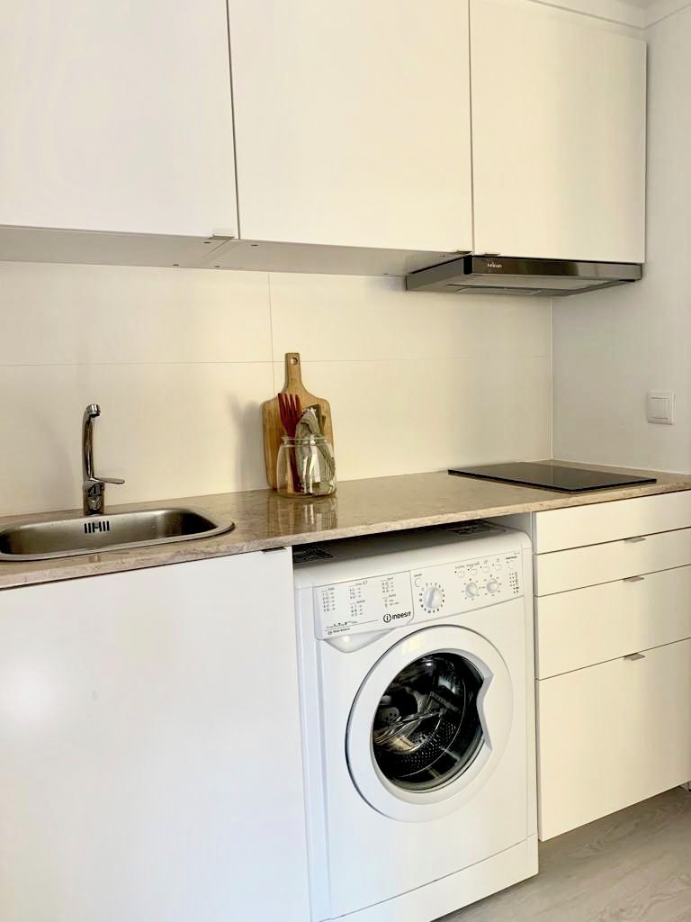 Een kleine keuken met een ingebouwde wasmachine, een roestvrijstalen spoelbak en een houten snijplank.
