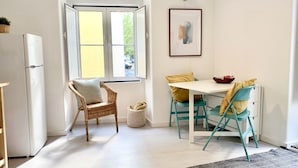 A compact kitchen with a refrigerator, a dining table with chairs, a window with curtains, and a framed picture on the wall.