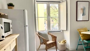 A kitchen with a white refrigerator, a wicker chair, a wicker basket, and a window with open shutters.