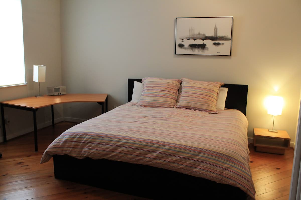 Un dormitorio ordenado con cama, mesitas de noche, una lámpara y un cuadro de un paisaje urbano en la pared.