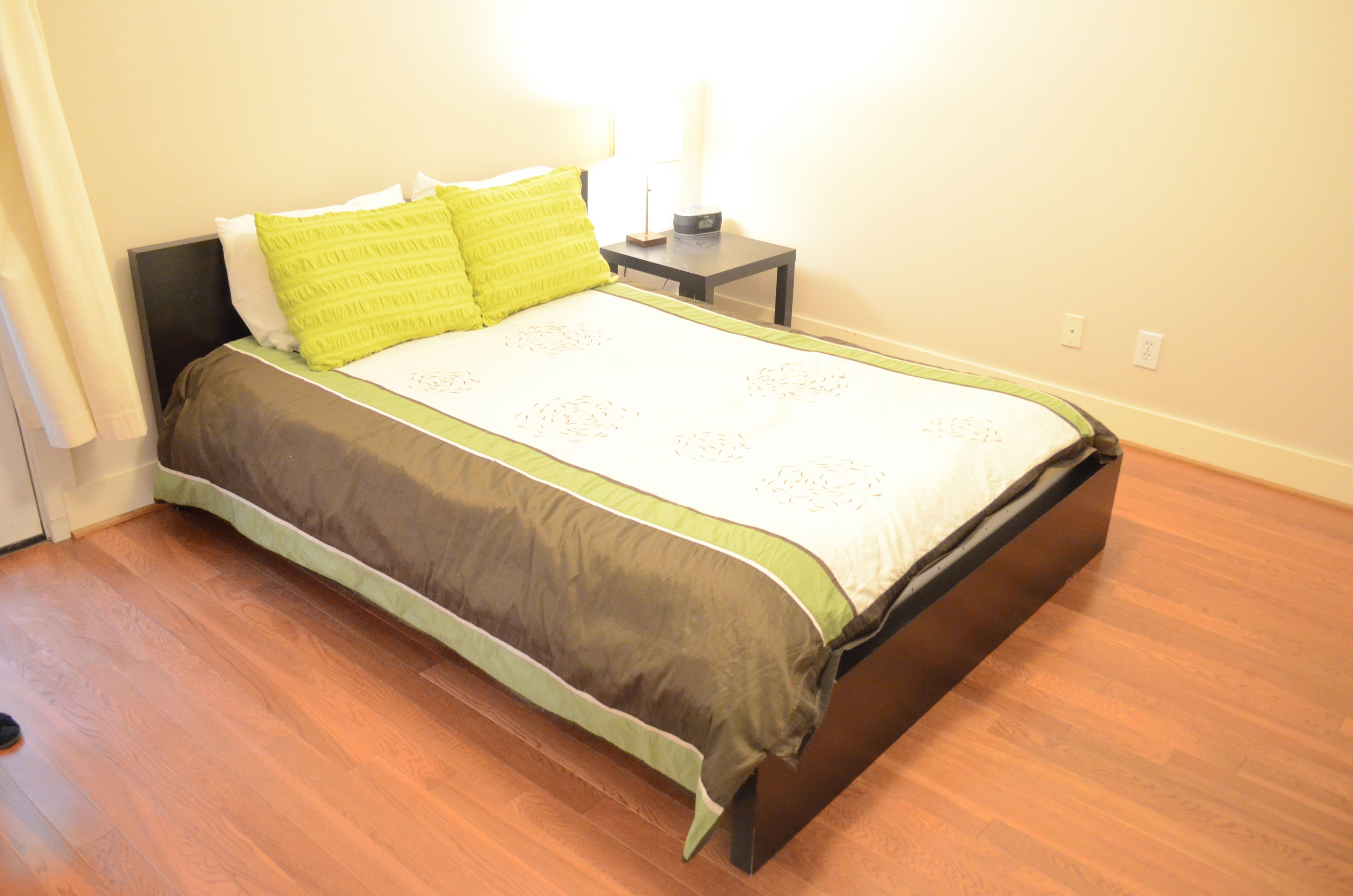 A neatly made bed with a green and white bedspread, a wooden floor, and a small bedside table with a lamp.