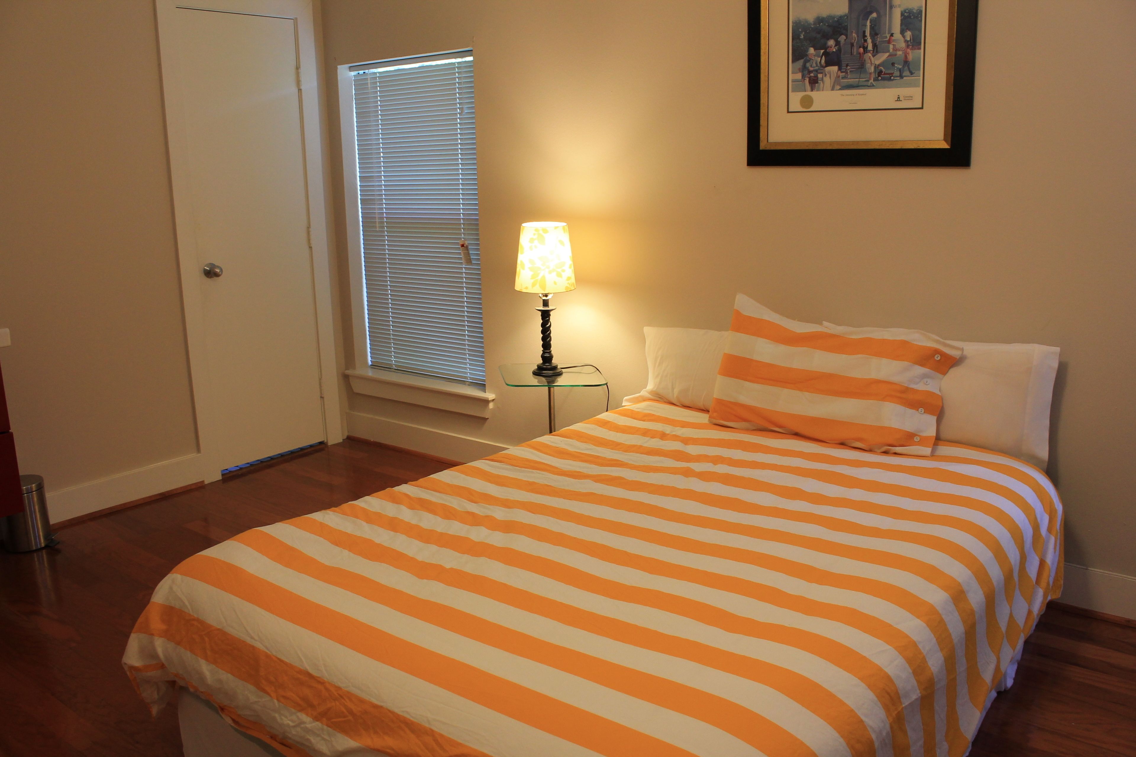 A bedroom with a bed, a nightstand, a lamp, a window with blinds, and a framed picture on the wall.