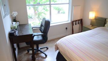 A bedroom with a bed, a desk with a chair, a window with a view of greenery, and a bedside lamp.