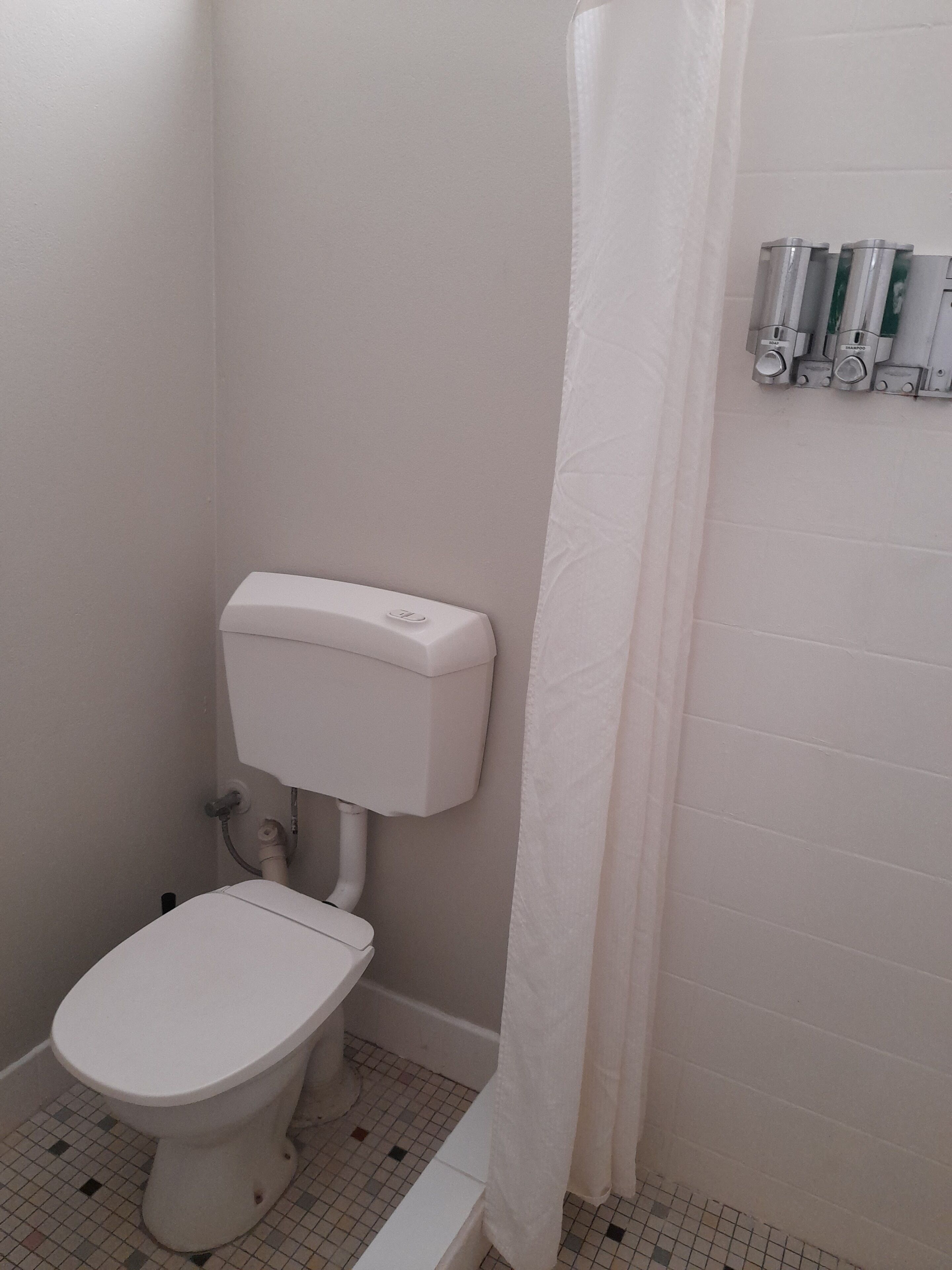 Un baño pequeño y blanco con inodoro, cortina de ducha y dispensador de jabón en la pared.