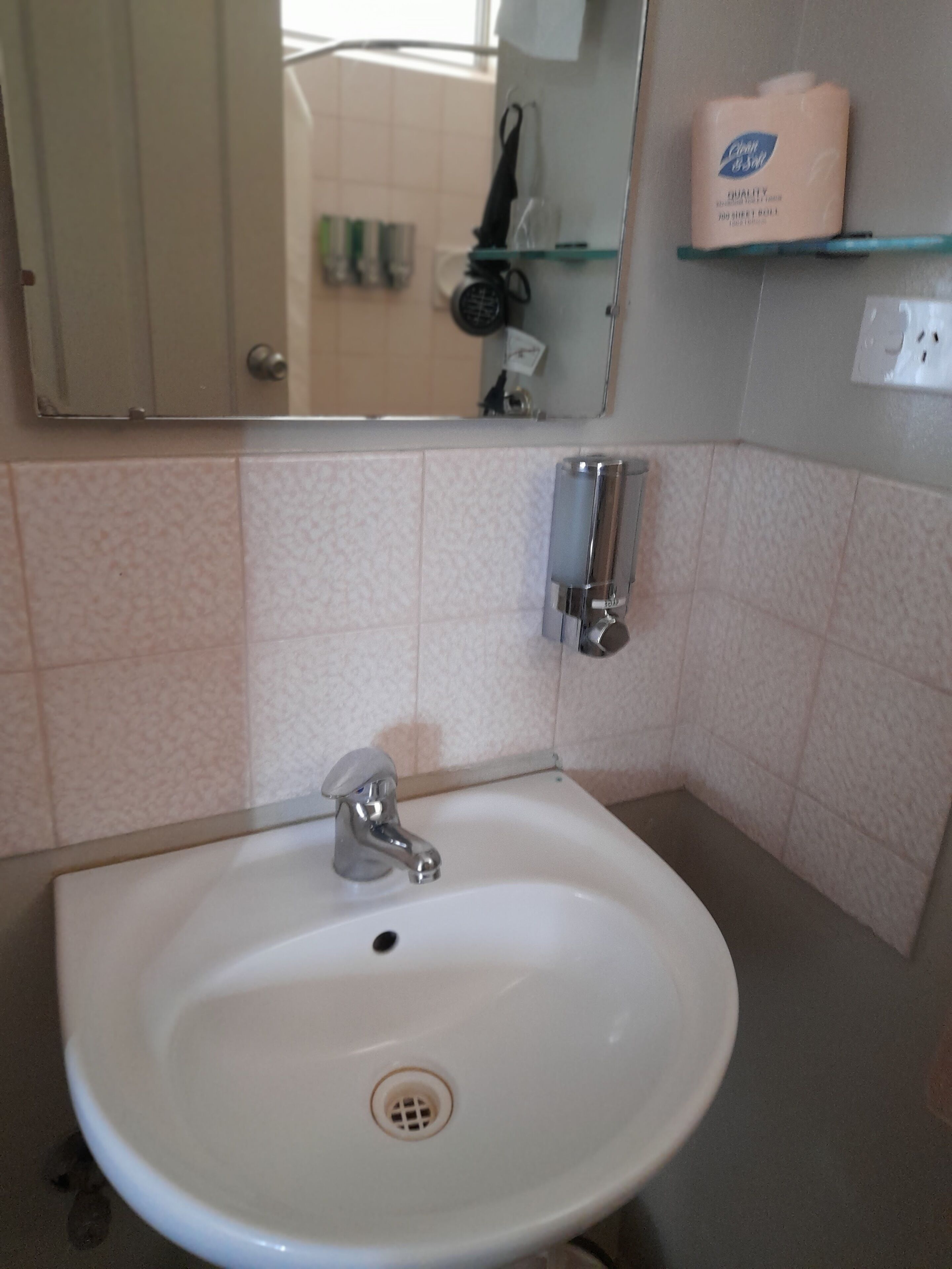 Un bany amb un lavabo blanc, un mirall i un dispensador de sabó sobre una estantàlia.