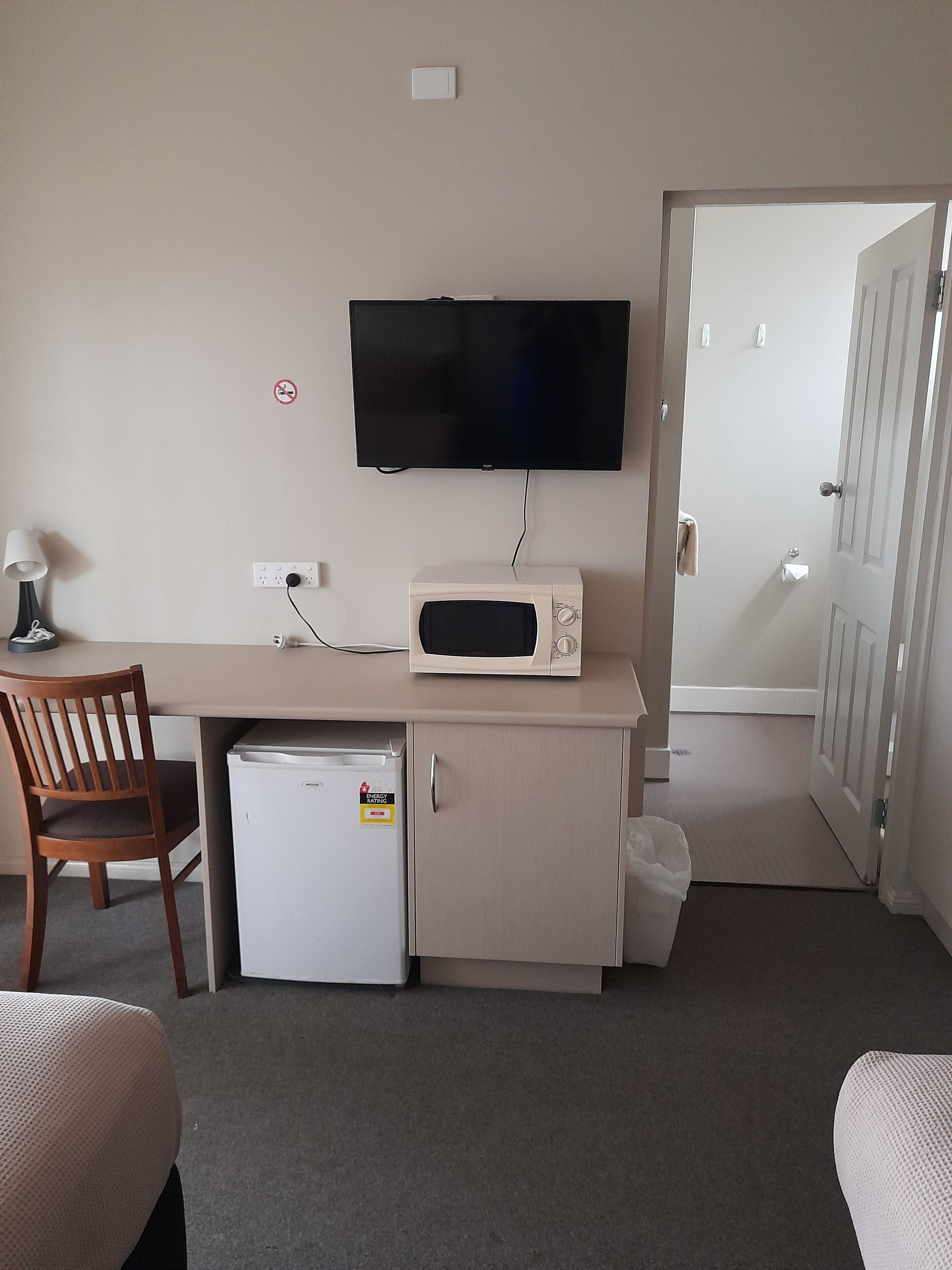 A compact hotel room with a kitchenette, a flat-screen TV mounted above the counter, a wooden chair, and a small refrigerator.
