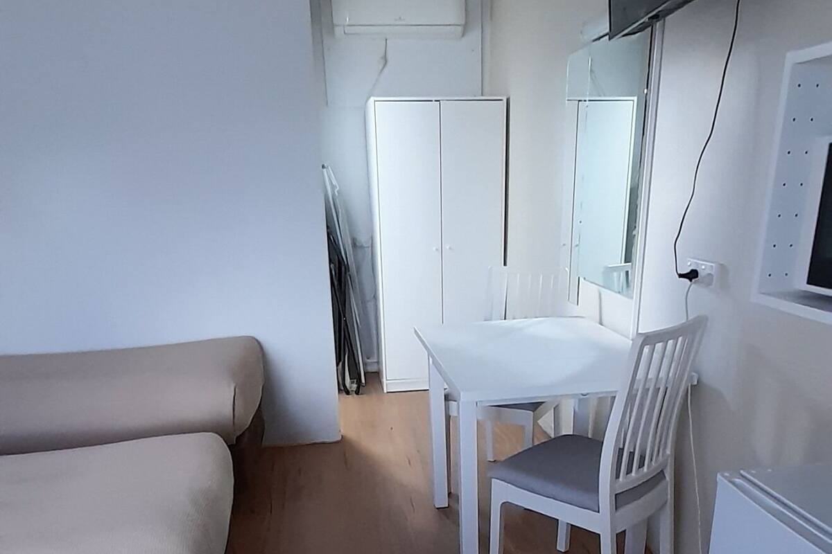 Huoneessa on sohva, ruokapöytä tuoleineen, seinälle asennettu televisio ja jääkaappi.