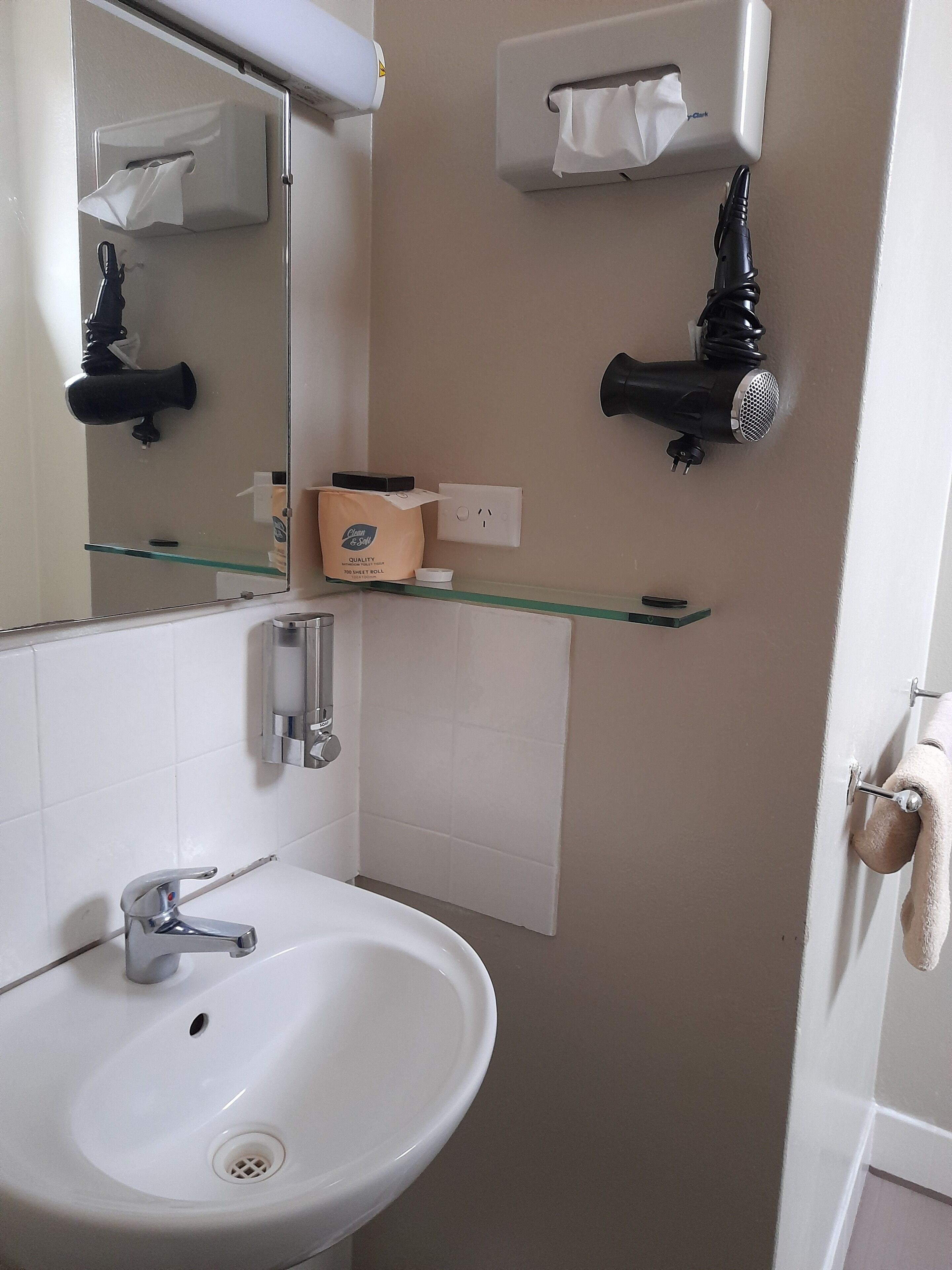 Un baño con un lavamanos blanco, un espejo, una repisa de vidrio con una caja y dos secadores de pelo montados en la pared.