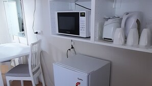 A compact room with a white desk, a small refrigerator, a microwave, and a shelf with white appliances.