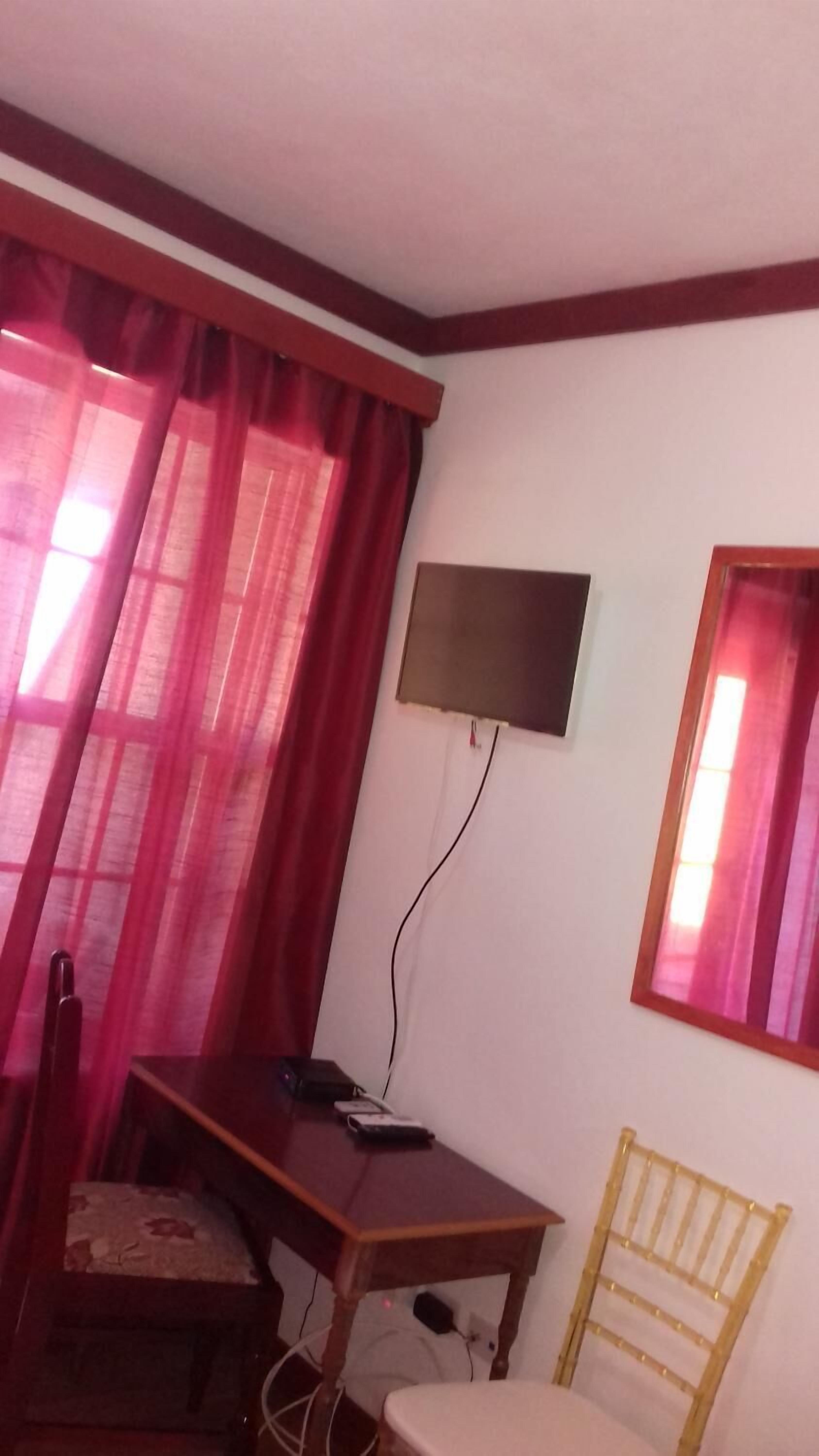A room with a wooden desk, a chair, a television mounted on the wall, and a window with curtains.