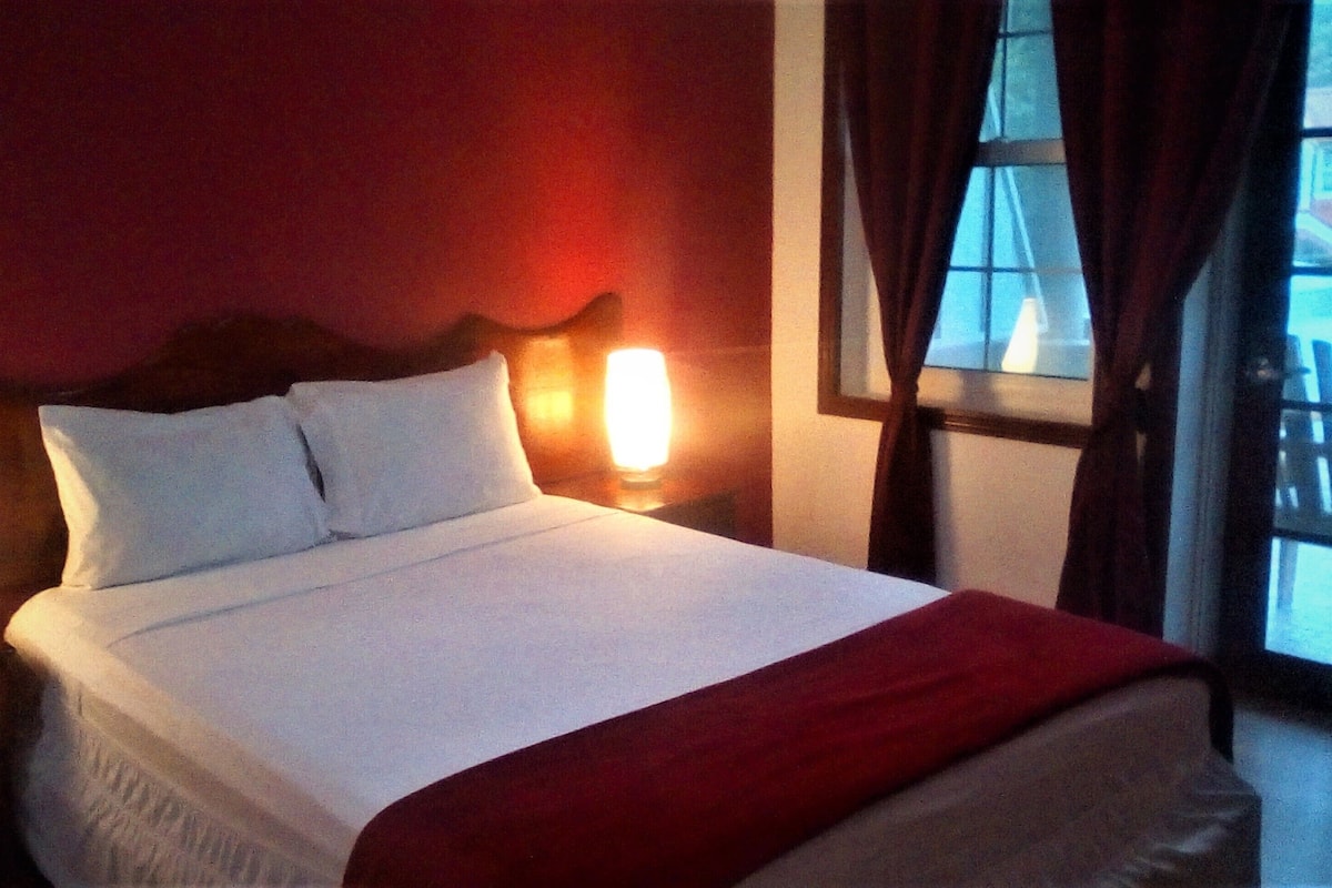 A neatly made bed with white linens and a red throw blanket, positioned in a room with red walls and a window with curtains.