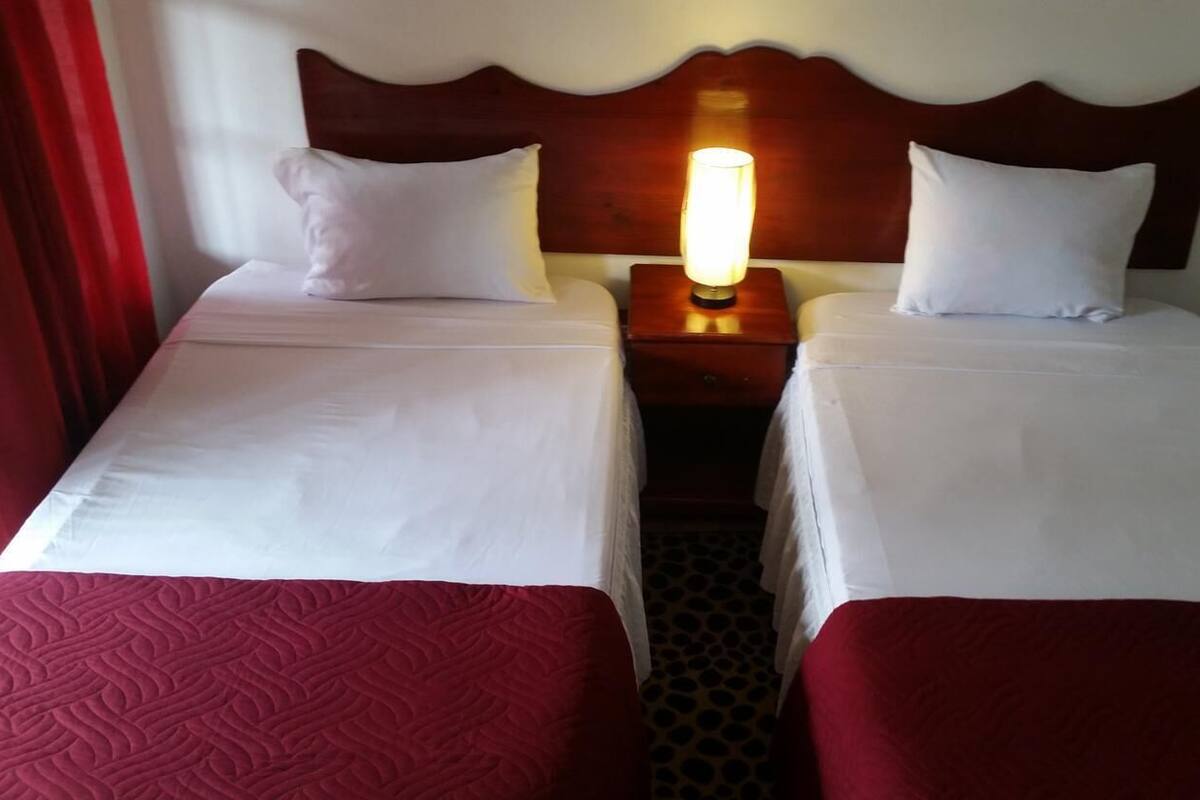 A hotel room with two beds, each with white linens and red bedspreads. There is a nightstand between the beds with a lamp on it.