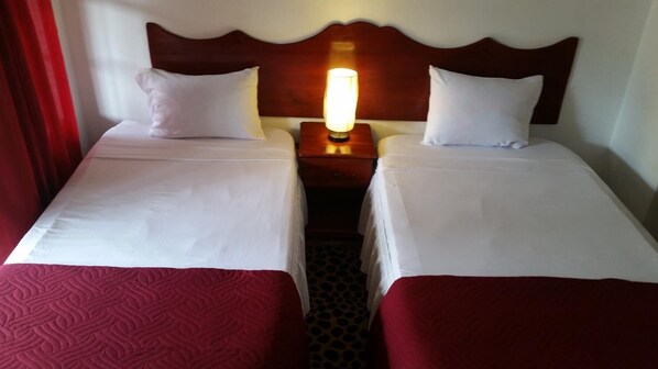 A hotel room with two beds, each with white linens and red bedspreads. There is a nightstand between the beds with a lamp on it.