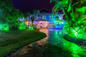 Una casa bien iluminada de dos pisos con un camino de piedra que lleva a una entrada con portón, rodeada de abundante vegetación.