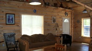 Interior acogedor de una cabaña con una mesa de comedor de madera, un sofá y una silla. Se ven ventanas que dejan entrar luz natural, una puerta y un techo con vigas a la vista.