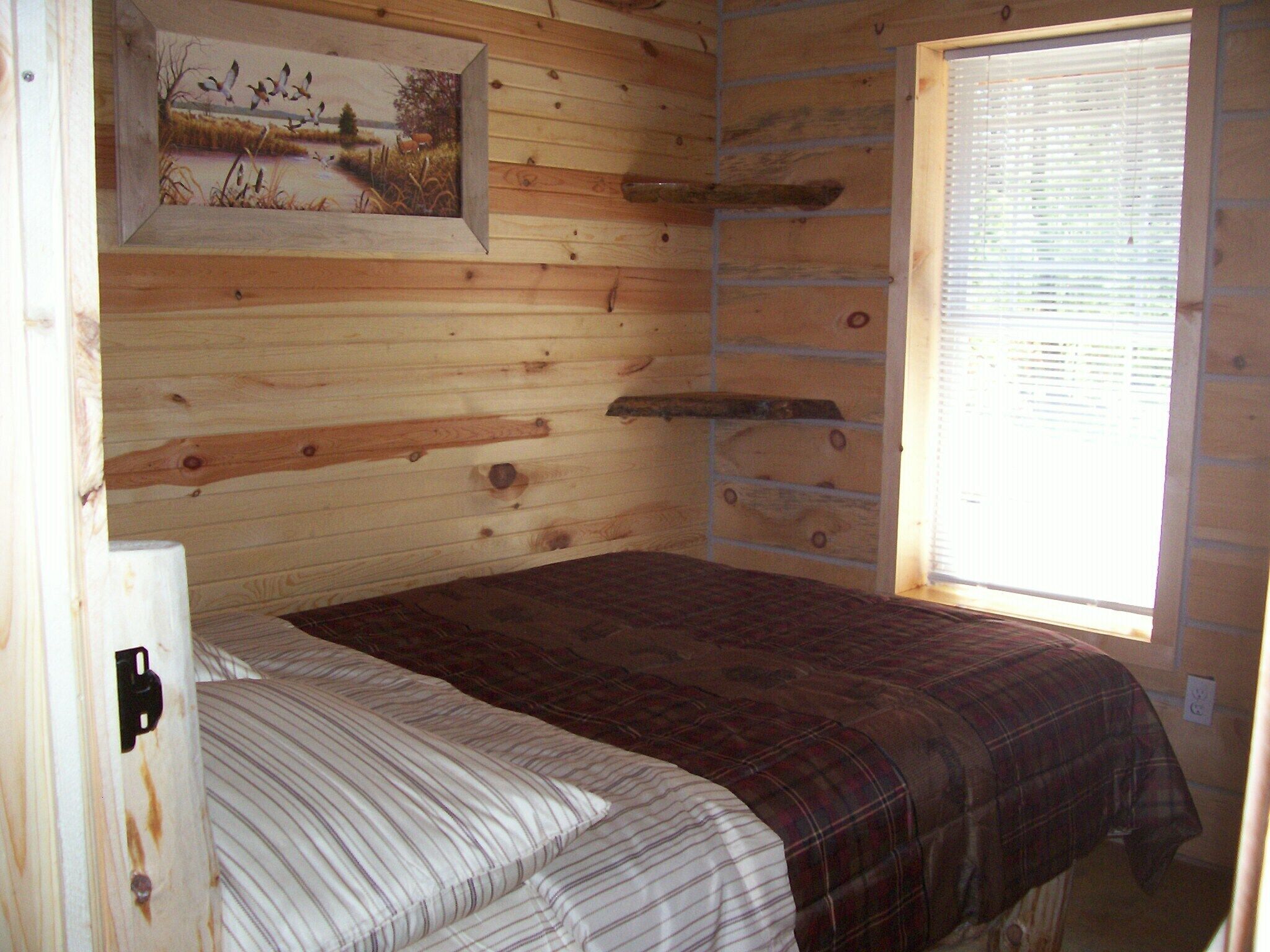 Une pièce de cabane en bois comprenant un lit, un tableau représentant des oiseaux et une fenêtre avec des stores.