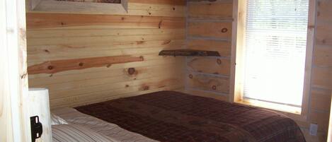 Une pièce de cabane en bois comprenant un lit, un tableau représentant des oiseaux et une fenêtre avec des stores.