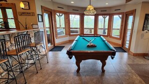 Une table de billard dans une pièce dotée de grandes fenêtres, un coin bar avec des tabourets et un porte-queue de billard.