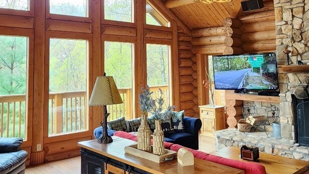 L’intérieur de cette vaste cabane en rondins est doté d’un grand plafond en bois, d’une cheminée en pierre, d’un téléviseur fixé au mur et d’une table à manger en bois, assortie de chaises à revêtement rouge.