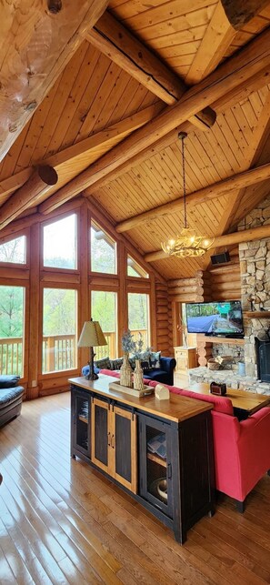 A spacious log cabin interior with a large wooden ceiling, a stone fireplace, a television mounted on the wall, and a wooden dining table with red upholstered chairs. - Log Home with Mountain view VERY PRIVATE!  Spa Sauna & Hot Tub "The Love Nest" (Maggie Valley)