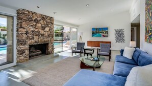 Ein modernes Wohnzimmer mit Kamin aus Stein, einem durch Glaswände sichtbaren Pool, einem blauen Sofa und einem Gemälde an der Wand.