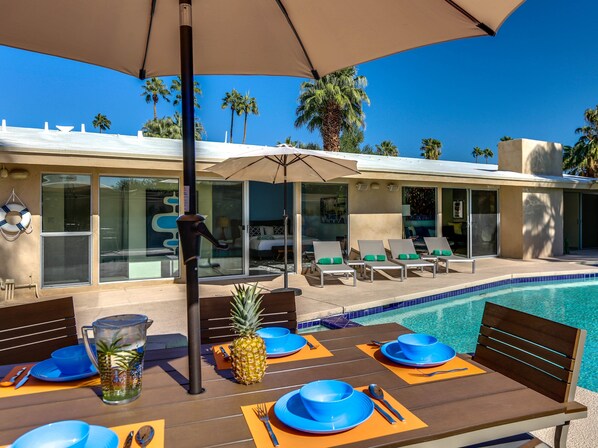 Zona de comedor junto a la piscina con una mesa para dos, una piña y una jarra de agua.