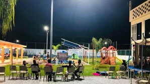 Ein Sitzbereich im Freien mit Stühlen und Tischen, ein Spielplatz mit Rutschen und Klettergerüsten sowie ein Gebäude mit großen Fenstern.