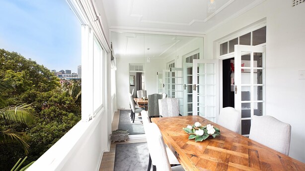 A modern dining area with a wooden table, white chairs, and a view of a cityscape and greenery.