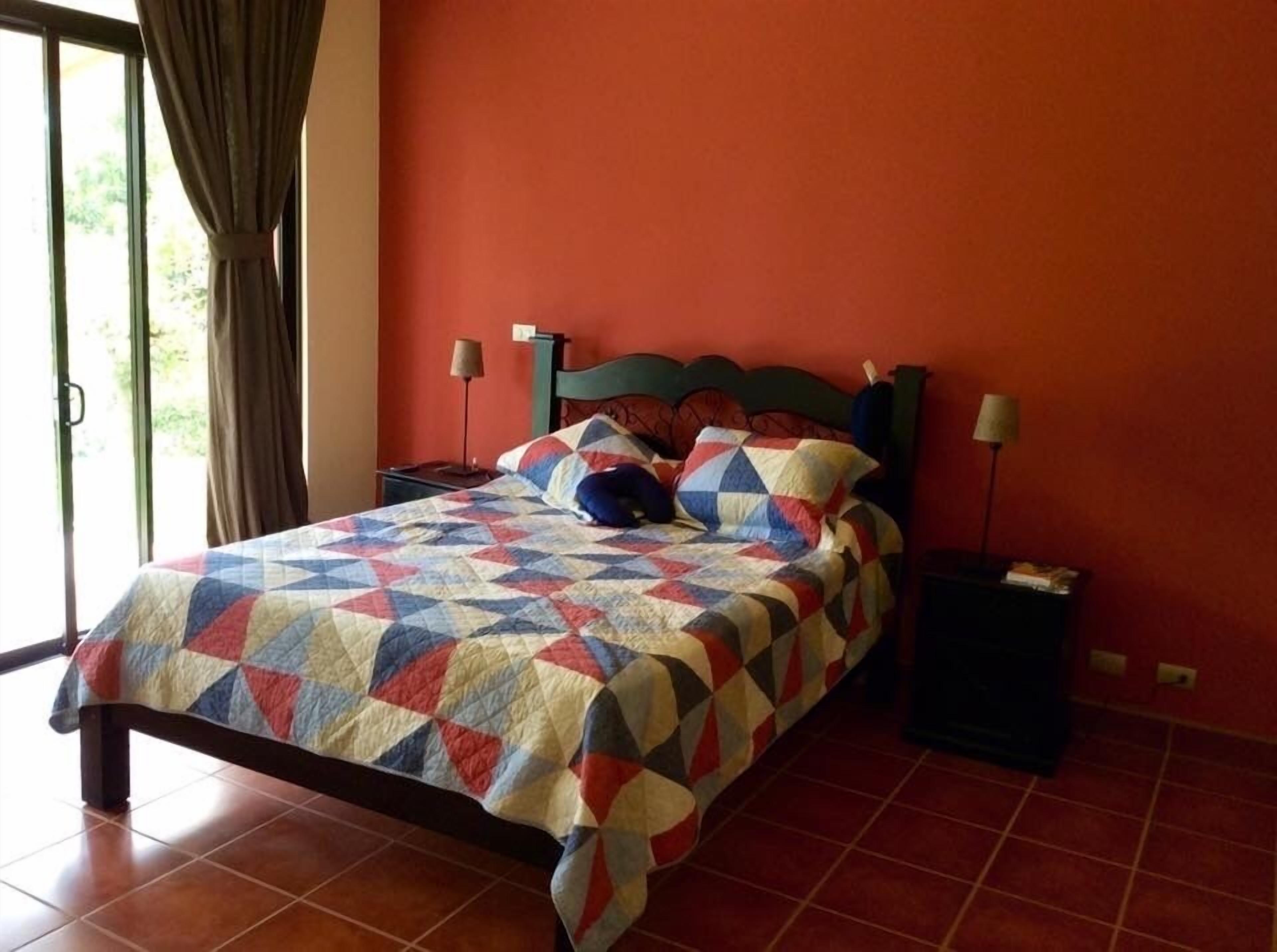 A bed with a colorful quilt, a nightstand with a lamp, and a window with curtains.