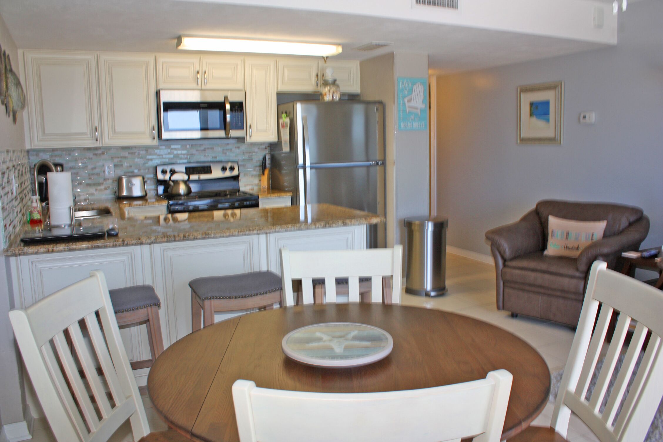 A kitchen with white cabinets, a stainless steel refrigerator, and a microwave. There is a dining table with four chairs.