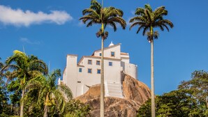 A large white building with multiple windows and a staircase leading up to the entrance, surrounded by palm trees and greenery.