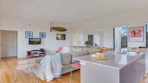 Ein modernes Wohnzimmer mit grauem Sofa, einem Couchtisch und einer Küchenzeile mit einer großen Kochinsel. An den Wänden hängen gerahmte Bilder und an der Küchenwand ein Poster.