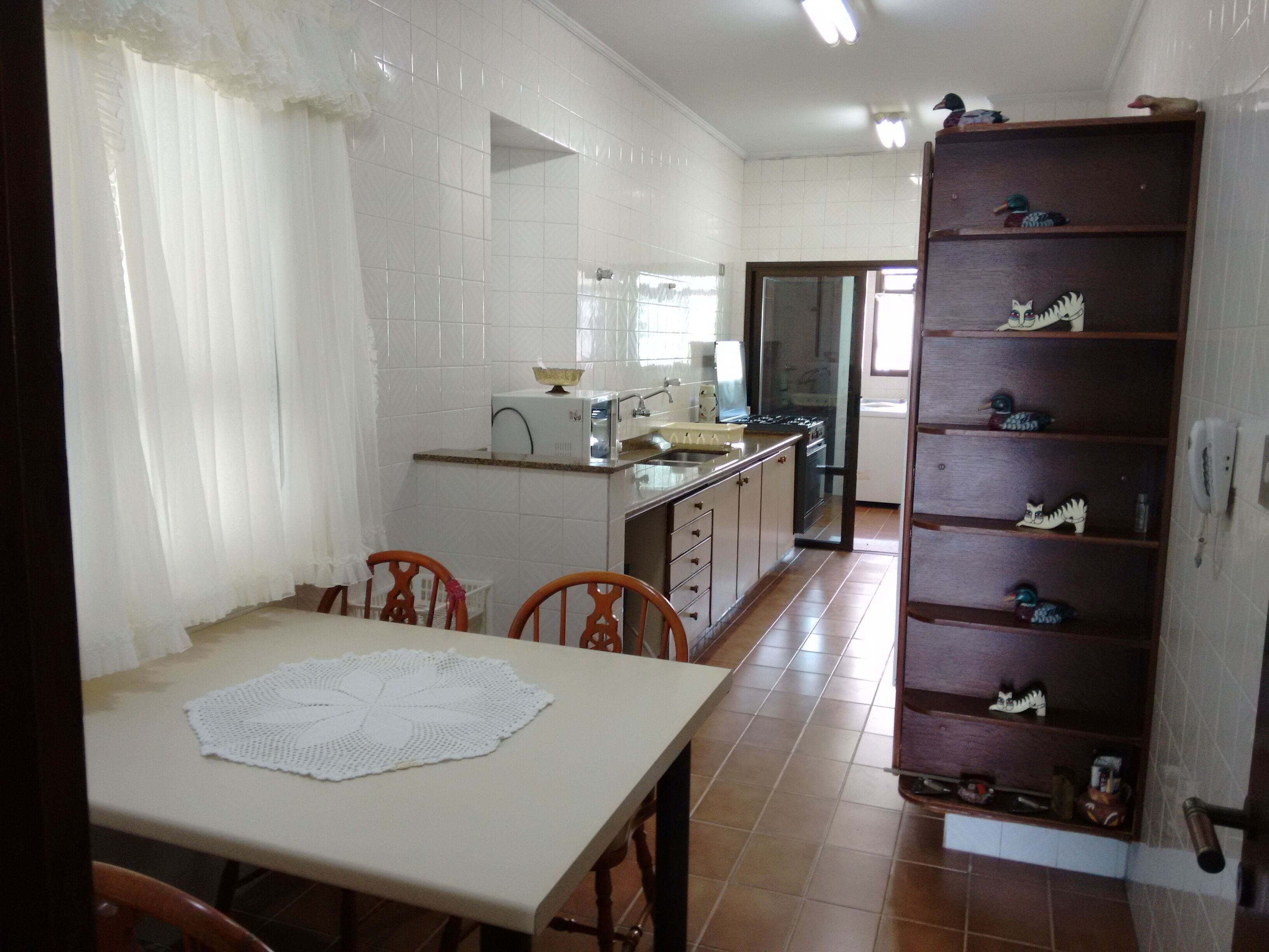 Une cuisine aux carreaux blancs, une table à manger recouverte d’une nappe blanche et une étagère en bois ornée d’objets décoratifs.