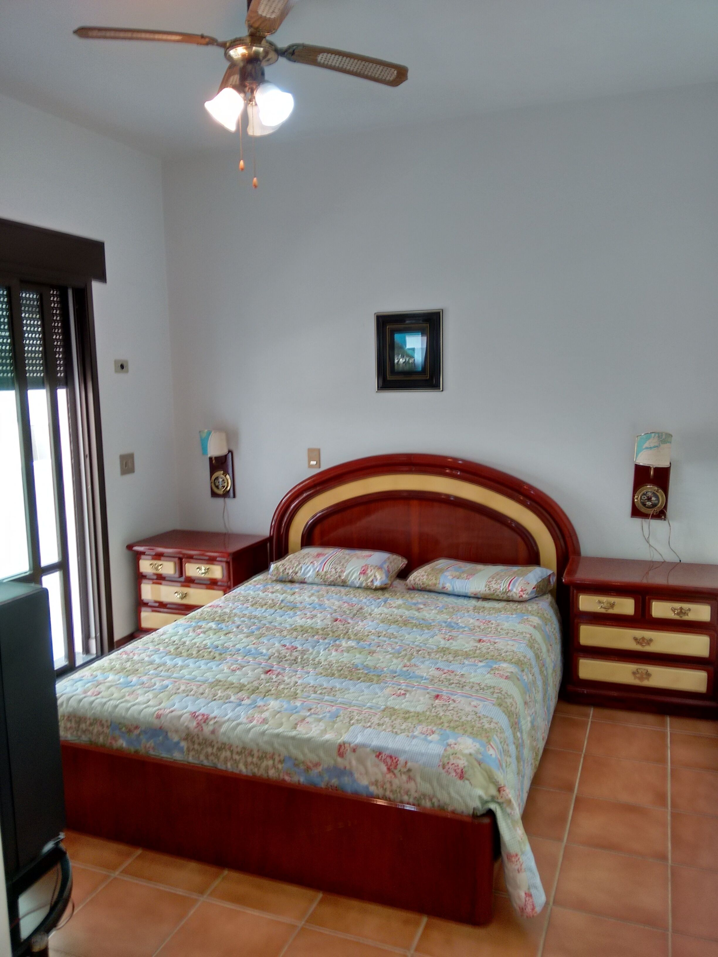 A bedroom with a wooden bed, two nightstands, a ceiling fan, a framed picture on the wall, and a window with blinds.