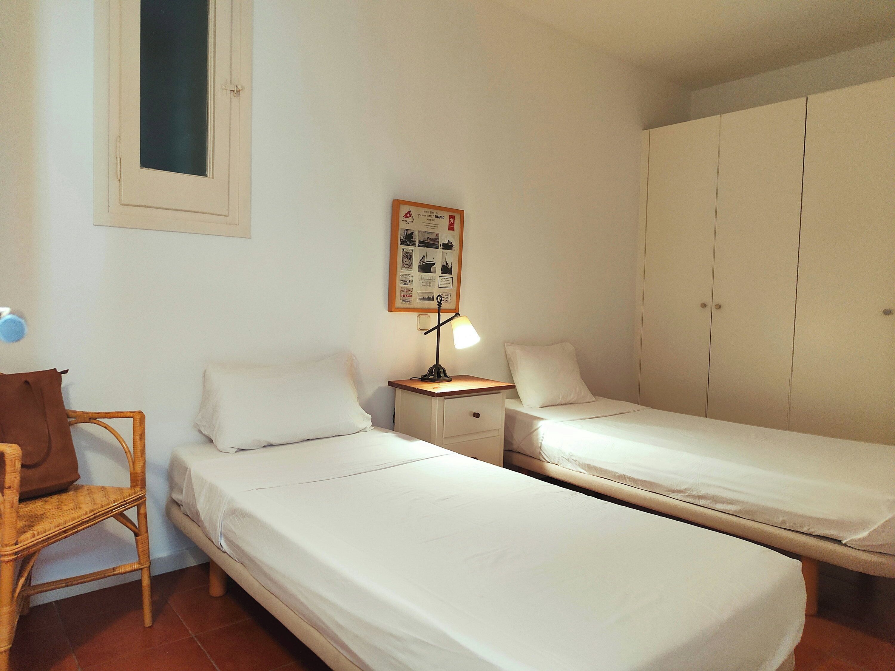 A room with two single beds, a nightstand with a lamp, a wicker chair, and a wardrobe.