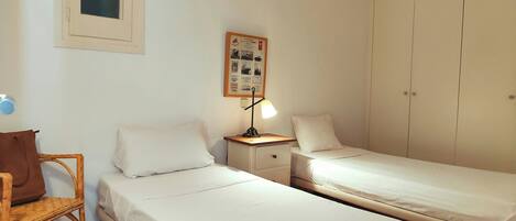 A room with two single beds, a nightstand with a lamp, a wicker chair, and a wardrobe.