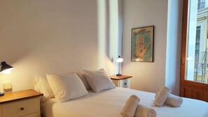 Ein ordentlich bezogenes Bett mit aufgerollten Handtüchern, einer Nachttischlampe, einem kleinen Nachttisch und einem gerahmten Poster an der Wand.