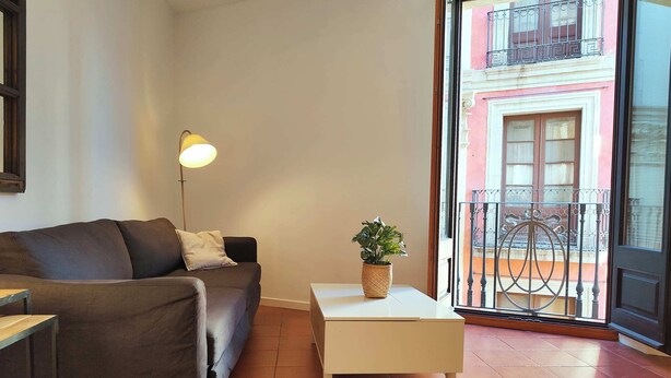 Ein Wohnzimmer mit einer Couch, einem kleinen Tisch mit einer Pflanze, einer Stehlampe und Blick auf einen Balkon mit Geländer.