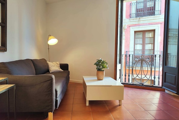 Ein Wohnzimmer mit einer Couch, einem kleinen Tisch mit einer Pflanze, einer Stehlampe und Blick auf einen Balkon mit Geländer.