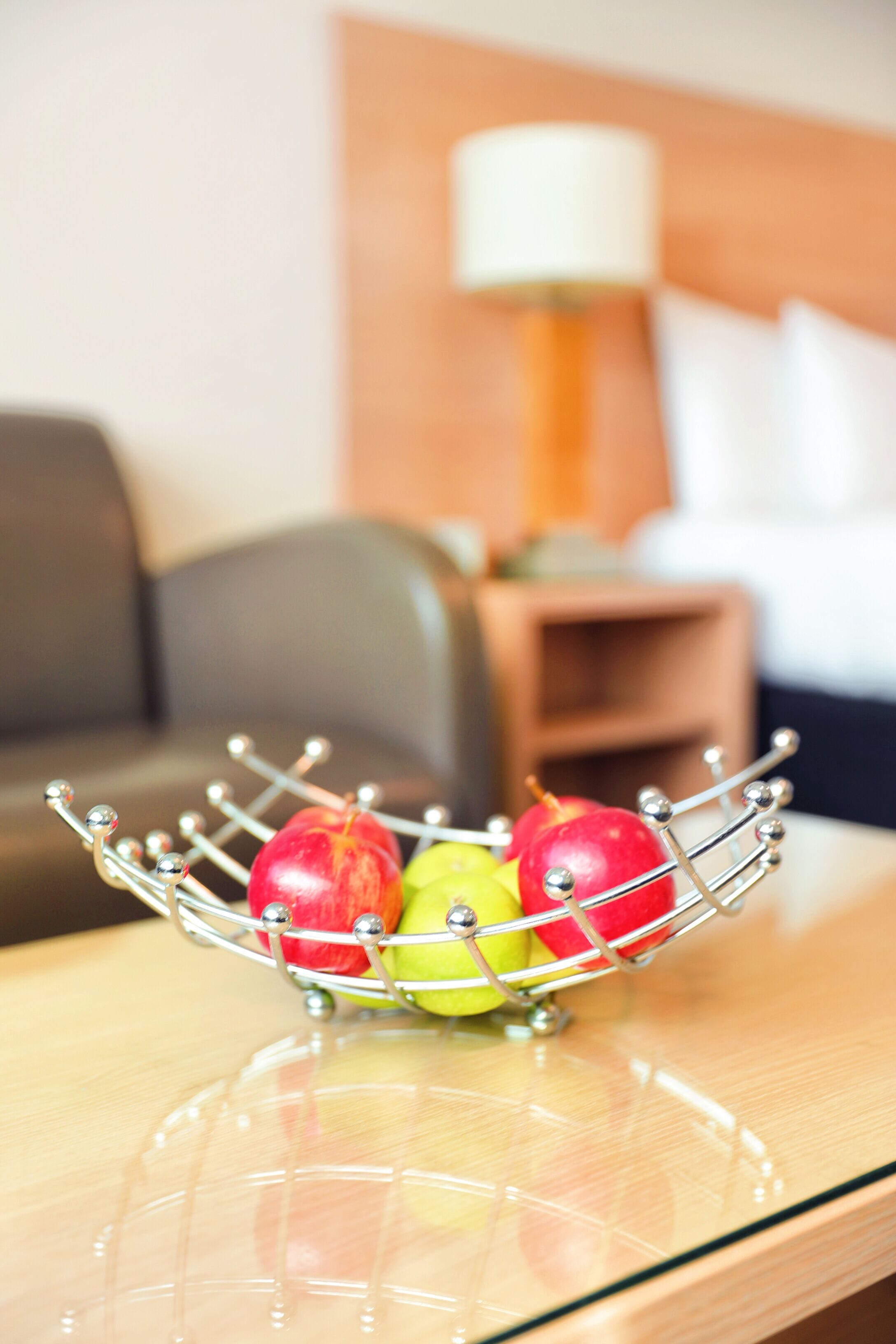A metal fruit basket with apples on a wooden table in a room with a sofa and a lamp in the background.