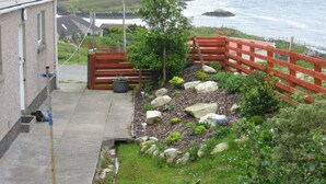 A coastal property with a paved pathway leading to a wooden gate, surrounded by a garden with various plants and rocks. - Shiantview Cottage - Magnificent Sea Views  (Stornoway, Isle of Lewis, Scotland)