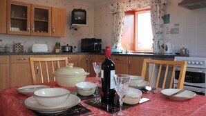 A kitchen with wooden cabinets, a microwave, a stove, and a dining table set with plates, bowls, and a wine bottle. - Shiantview Cottage - Magnificent Sea Views  (Stornoway, Isle of Lewis, Scotland)