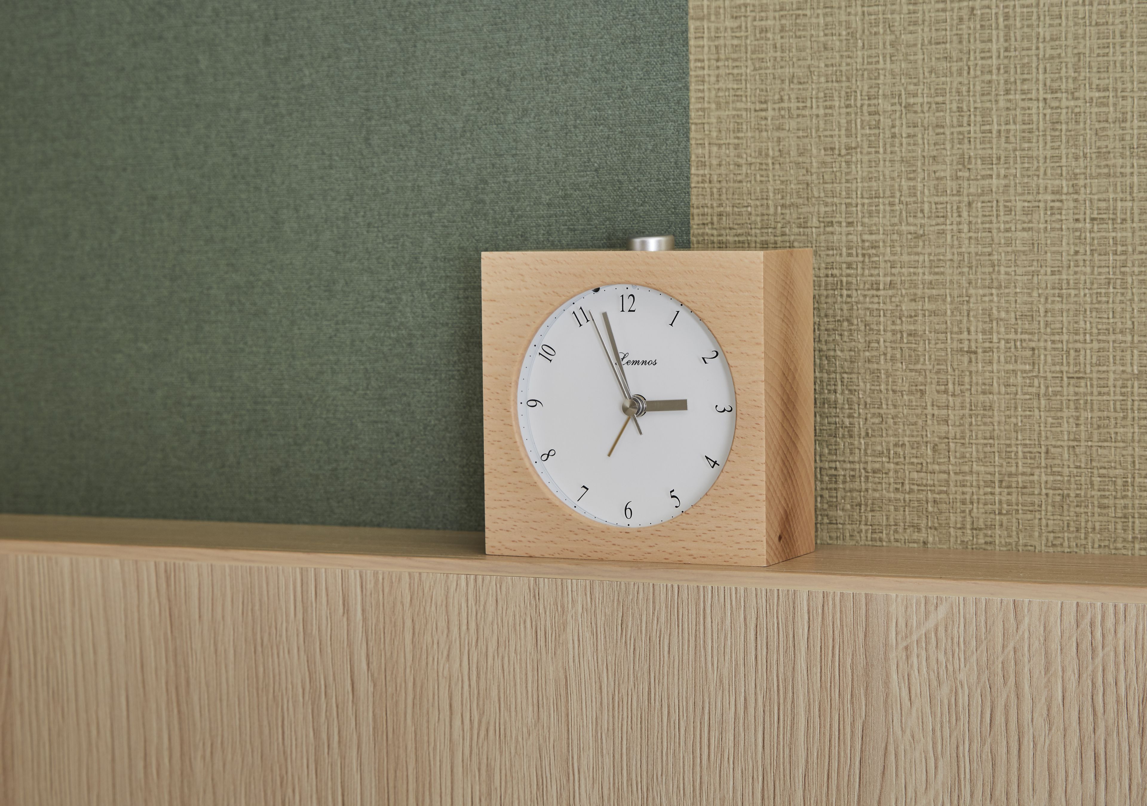 A wooden clock with a white face and black numbers, placed on a wooden shelf against a green and beige textured wall.