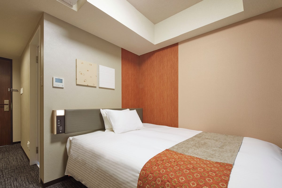 A hotel room with a bed, a control panel, a thermostat, and a door.