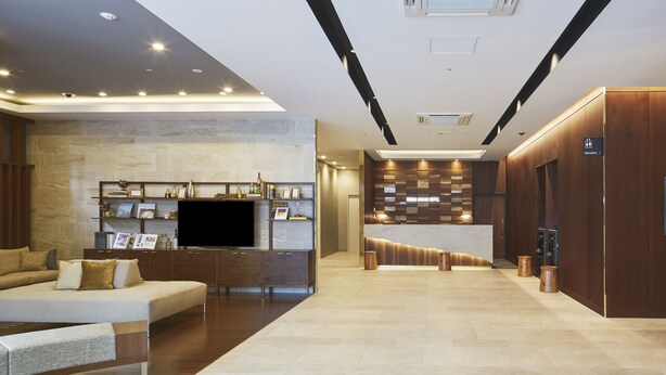 A modern hotel lobby with a sofa, a flat-screen TV, a reception desk, and a wooden reception area. - Iroha Grand Hotel (Matsumoto)