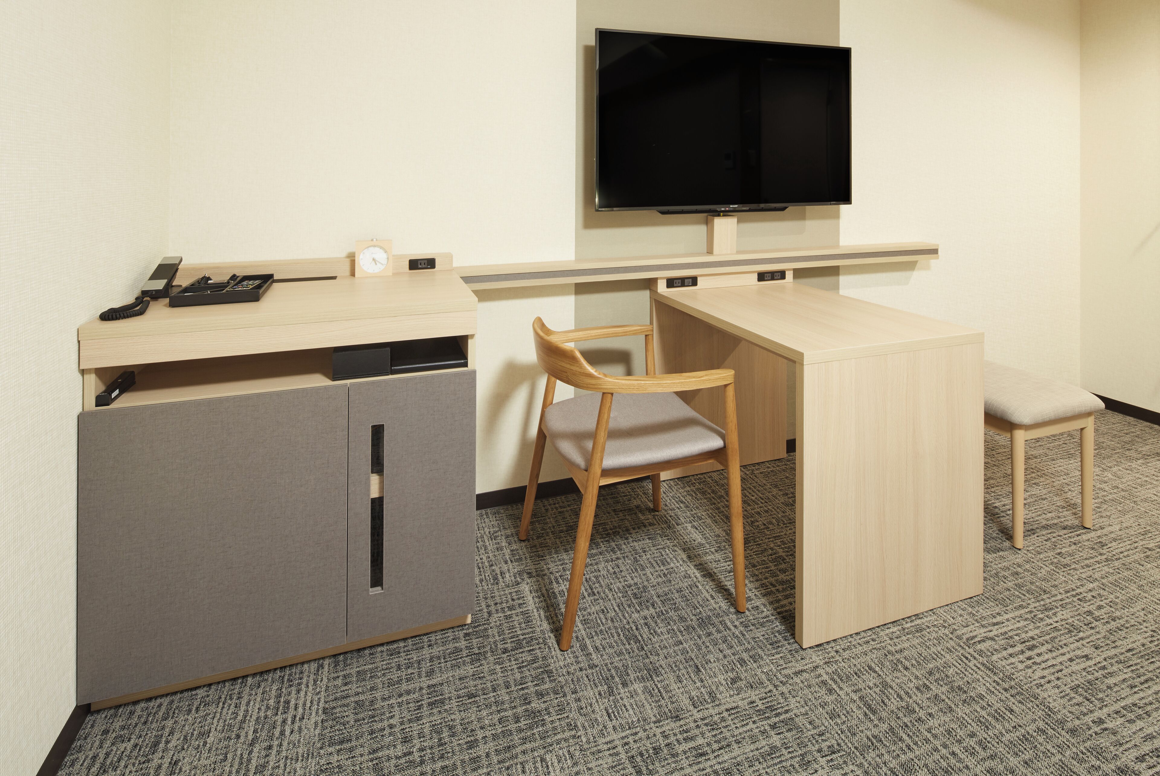A small, compact hotel room with a single bed, a desk with a chair, a television mounted on the wall, and a small cabinet.