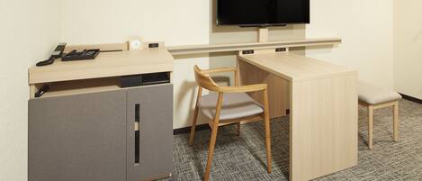 A small, compact hotel room with a single bed, a desk with a chair, a television mounted on the wall, and a small cabinet.