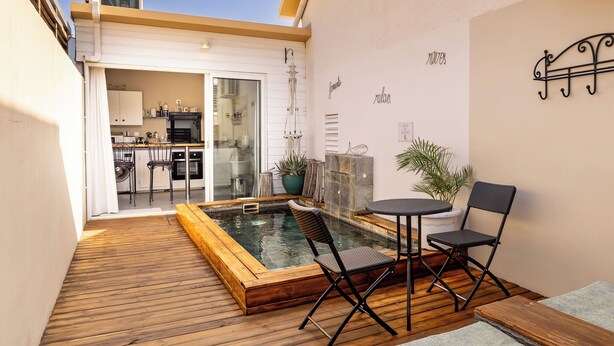 Un terrazzo in legno con una vasca idromassaggio, due sedie e un tavolo. Si intravede una cucina attraverso una porta aperta.