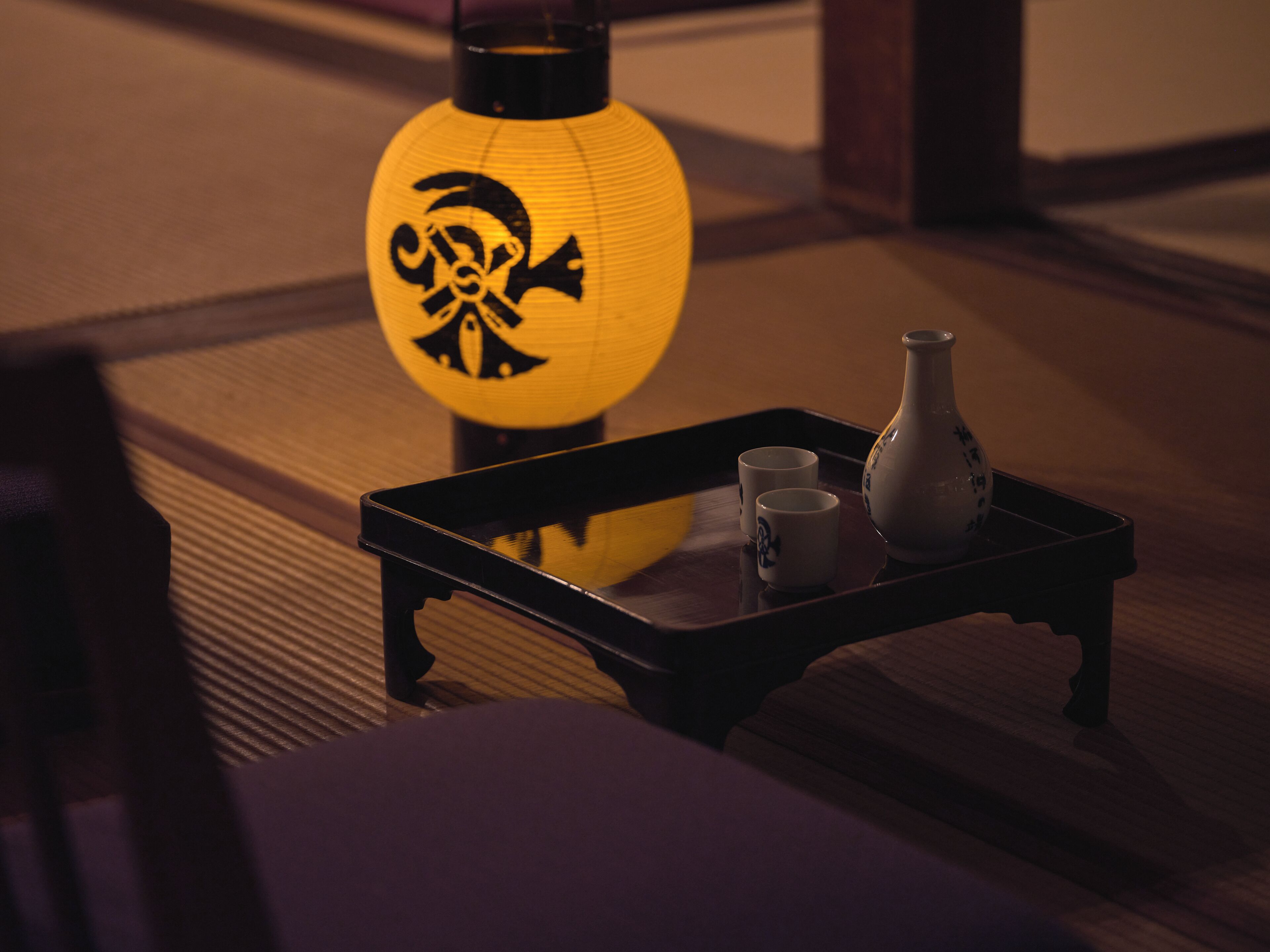 A traditional Japanese room with tatami flooring, a black lacquered table with two cups and a vase, and a yellow lantern with a black design.